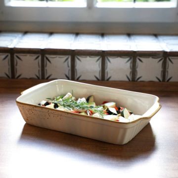 Rechteckige, weiße Keramik-Auflaufform mit leicht braunem Rand, gefüllt mit geschnittenem Gemüse wie Zucchini und Paprika, dekoriert mit Rosmarinzweigen. Die Form steht auf einer hölzernen Oberfläche vor einem gemusterten Hintergrund.