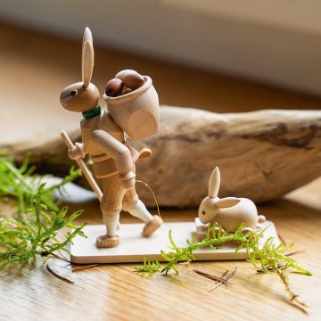 Holzfigur eines gehenden Hasen mit Rucksack voller Eier und Wanderstock, führt auf einer rechteckigen Bodenplatte einen kleinen Hasen auf einem Schlitten; Dekoration mit Moos und Holzstück im Hintergrund.