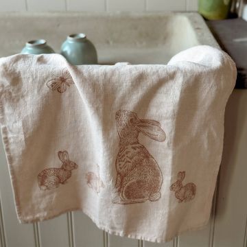 Ein cremefarbenes Küchentuch mit braunen Zeichnungen von Hasen und einem Schmetterling hängt über einem Wannenrand. Im Hintergrund sind unscharf blaue Vasen zu sehen.