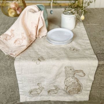 Tischläufer und Geschirrtuch aus Leinen mit aufgedruckten Hasen und Schmetterlingen, dekorativ auf einem gedeckten Tisch mit Tellern, Tassen und Vase arrangiert. Im Hintergrund Osterdekoration mit Eiern in einem Drahtkorb.