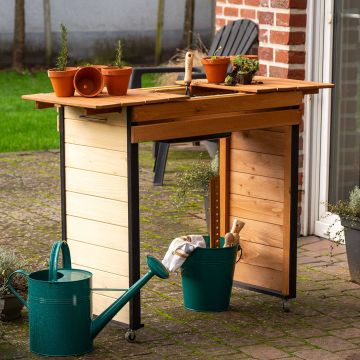 Ein hölzerner Pflanztisch auf einer Terrasse mit Blumentöpfen und Gartengeräten. Daneben steht eine grüne Gießkanne und ein Eimer mit Werkzeug. Der Tisch hat Räder und ist teilweise klappbar.