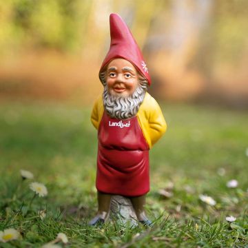 Ein Gartenzwerg mit rotem Spitzhut und roter Schürze, auf der "Landlust" steht. Der Zwerg hat grauen Bart, gelbes Hemd und steht auf einem grünen Rasen mit Gänseblümchen im Hintergrund.