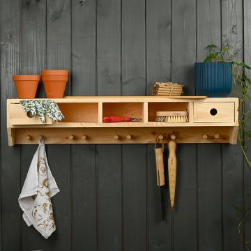 Ein hölzernes Wandregal mit mehreren Fächern, Tongefäßen, einer Schere, Bürste, Pflanzstöcken und einem blauen Blumentopf. Darunter befinden sich Haken mit aufgehängten Gartenhandschuhen und Stofftüchern vor einer dunkelgrauen Holzwand.