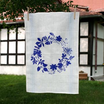 Weißes Geschirrtuch mit einem blauen Blumenkranzmotiv hängt an einer Leine im Freien. Im Hintergrund sind ein Fachwerkgebäude und grüne Bäume sichtbar.