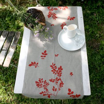 Grauer Tischläufer mit roten Blättermotiven liegt auf einem weißen Holztisch im Freien. Auf dem Tisch stehen eine weiße Kanne, eine Glasschale und ein Korb mit Blumen. Daneben steht eine Holzbank.