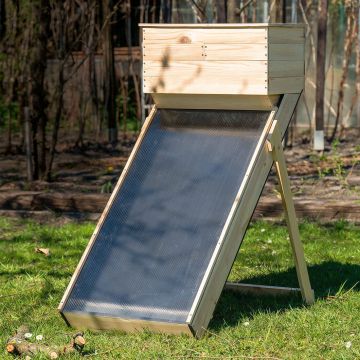 Holzgestell mit geneigtem Solarpanel im Garten aufgestellt. Im Hintergrund sind Bäume und Büsche zu sehen. Das Solarpanel ist schwarz und wird von Holzrahmen gestützt.