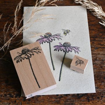 Zwei Holzstempel mit Illustrationen einer Blume und einer Biene liegen auf Papier mit gedruckten Blumen und einer Biene. Trockene Grashalme sind als Dekoration arrangiert.