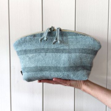 Hand hält eine gewebte, hellblaue Kosmetiktasche mit Reißverschluss und Kordelzug, vor einer weißen, vertikalen Holzverkleidung. Die Tasche hat horizontale Streifenmuster und liegt mittig auf der Handfläche.