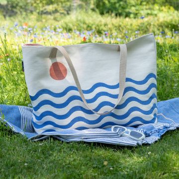 Große Stofftasche mit blauen Wellen und rotem Kreis darauf, steht auf einer blauen Decke, die auf Gras liegt. Eine Sonnenbrille ist auf der Decke platziert. Im Hintergrund ist eine blühende Wiese zu sehen.