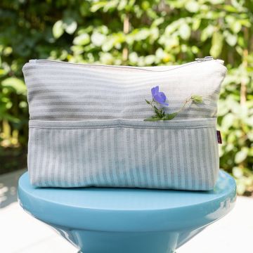 Hellgraue gestreifte Stofftasche mit Reißverschluss, auf einem blauen Tisch im Freien platziert. Eine kleine blaue Blume mit Blättern steckt in der vorderen Tasche der Tasche. Hintergrund unscharf grün.