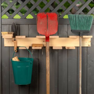 Gartengeräte hängen an einer hellen Holzleiste vor einem dunklen Holzzaun. Sichtbar sind ein Spaten, ein Besen, eine kleine Schaufel, eine Bürste und ein grüner Eimer mit Griff. Im Hintergrund ein gitterartiger Zaunabschluss.