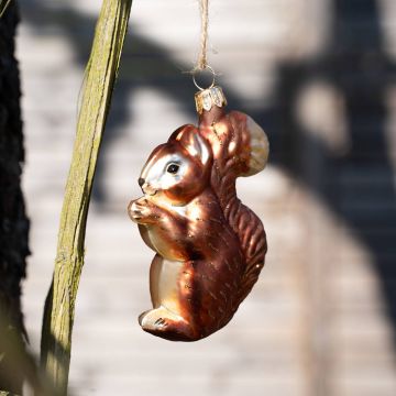 Glasornament in Form eines rotbraunen Eichhörnchens mit goldenen Details, hängend an einem Ast vor unscharfem Hintergrund.