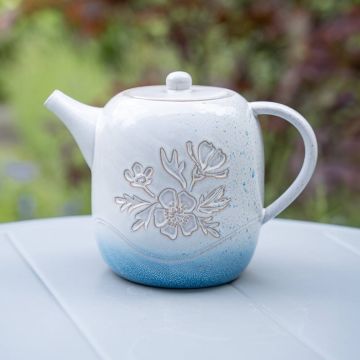 Eine weiße Teekanne mit blauen Sprenkeln, verziert mit einem floralen Gravurmuster, steht auf einem grauen Tisch. Die Kanne hat einen seitlichen Henkel und eine Ausgusstülle.