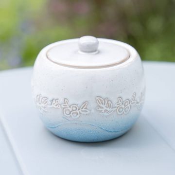 Weißer Keramik-Zuckerdose mit blauem Farbverlauf und floraler Gravur. Mit Deckel versehen. Steht auf einem grauen Tisch mit einem unscharfen, grünen Hintergrund.