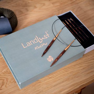 Ein Set von Rundstricknadeln auf einer geöffneten „Landlust KnitPro“-Box liegt auf einem Holztisch. Daneben befindet sich ein dunkelgrünes Wollknäuel.
