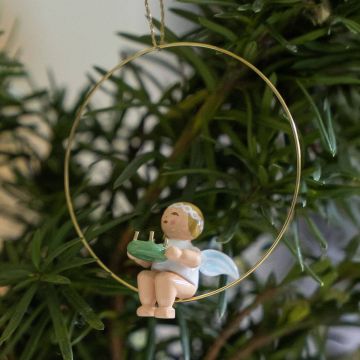 Ein kleiner Engel aus Holz sitzt in einem goldenen Metallring und hält ein Musikinstrument. Der Engel trägt Flügel und hat ein besetztes Kopftuch. Im Hintergrund sind grüne Pflanzen zu sehen.