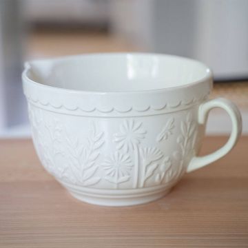 Weiße Keramik-Rührschüssel mit floralem Reliefmuster und einem Ausgießer. Die Schüssel hat einen Henkel und steht auf einer Holztischoberfläche. Der Hintergrund ist unscharf.