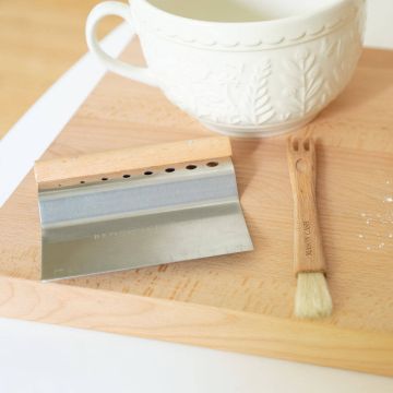 Holzschneidebrett mit einem Metall-Teigschaber und einem Backpinsel aus Holz. Eine große, verzierte, weiße Keramiktasse steht im Hintergrund. Der Teigschaber liegt flach, der Pinsel zeigt zur Bildkante.