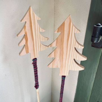 Zwei Holzdekorationen in Tannenbaumform auf Stäben, mit bunter Wolle umwickelt. Die Dekorationen lehnen an einer Wand neben einer grün gestrichenen Tür.