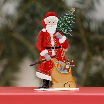 Ein Weihnachtsmann aus Zinn in rotem Anzug hält einen geschmückten Tannenbaum. Neben ihm steht ein Sack voller Spielzeug, darunter ein Teddybär und ein Ball. Die Figur steht auf einem roten Untergrund vor verschwommenem Hintergrund.
