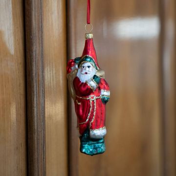 Handbemalter Weihnachtsmann-Anhänger aus Glas in rotem Mantel und Mütze, mit goldenem Sack auf dem Rücken, an rotem Band vor holzfarbener Wand hängend.