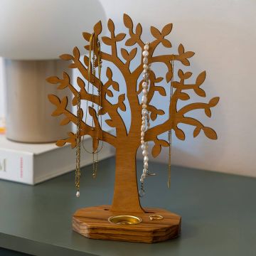 Holz-Schmuckständer in Form eines stilisierten Baumes mit vielen Ästen und Blättern, auf dessen Zweigen Ketten und Schmuck hängen; im Holzsockel befindet sich eine kleine runde Ablage für Ringe, auf einer Kommode vor Lampe und Buch platziert.