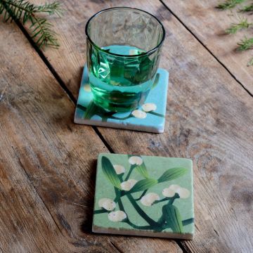 Zwei quadratische Keramikuntersetzer mit grün-blauem Hintergrund und Mistelzweig-Motiv aus grünen Blättern und weißen Beeren auf einer rustikalen Holzoberfläche, einer mit einem grünen Wasserglas, der andere leer im Vordergrund platziert.