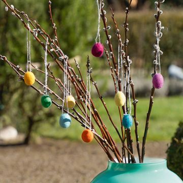Bunte Filzeier hängen an braunen Zweigen in einer türkisfarbenen Vase, weicher Filz, frische Frühlingsfarben.
