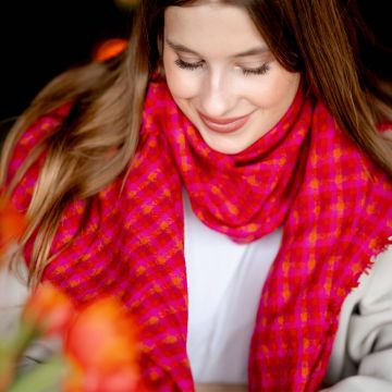 Frau mit langen braunen Haaren trägt leuchtend pink karierten Schal, sanfte rote und orange Töne, weiche Stofftextur.