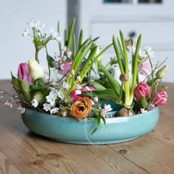 Runder, türkisfarbener Keramikkranz mit bunten Frühlingsblumen, grünen Blättern und Knollen auf hellem Holztisch.