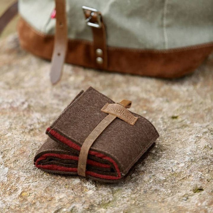 Kompakte braune Filztasche mit Lederband, auf einer Steinoberfläche liegend, in der Nähe eines grün-braunen Rucksacks. Die Tasche ist mit einem Etikett versehen, auf dem "Landlust" steht.