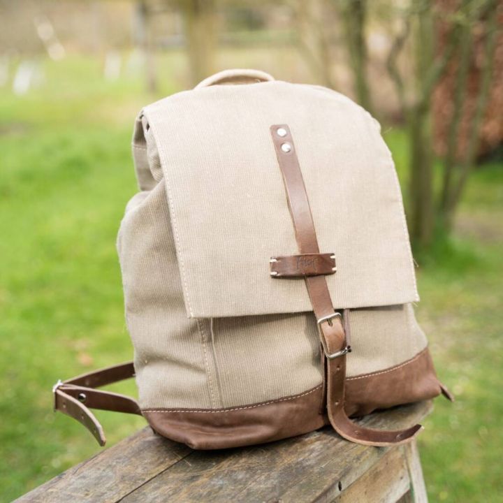Beiger Stoffrucksack mit braunen Lederdetails und Schnallen, auf einem Holztisch im Freien. Der Rucksack hat eine Klappe mit Lederriemenverschluss. Im Hintergrund unscharf grüne Wiese und Bäume.