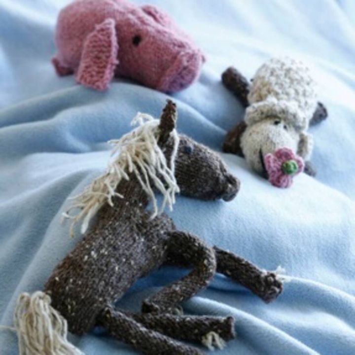 Drei gestrickte Stofftiere liegen auf einer hellblauen Decke: ein rosa Schwein oben links, ein weißes Schaf mit rosa Blume im Maul oben rechts und ein braunes Pferd mit heller Mähne und Schweif unten im Vordergrund. Alle Tiere bestehen aus Wolle und haben