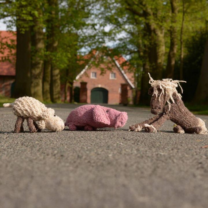 Drei gestrickte Stofftiere liegen auf einer hellblauen Decke: ein rosa Schwein oben links, ein wei&szlig;es Schaf mit rosa Blume im Maul oben rechts und ein braunes Pferd mit heller M&auml;hne und Schweif unten im Vordergrund. Alle Tiere bestehen aus Wolle und haben
