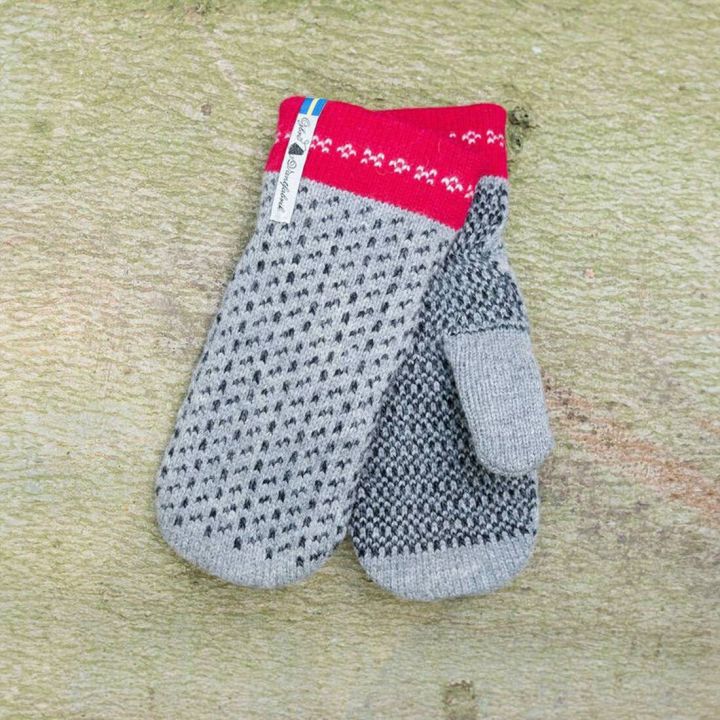Ein Paar gestrickte Fäustlinge liegt auf hellem Untergrund; sie sind grau mit schwarzem Muster, rotem Bündchen mit weißem Muster und unterschiedlich gestalteten Handflächen, die rechte mit dunklerem Muster. Ein Etikett ist am Bündchen befestigt.