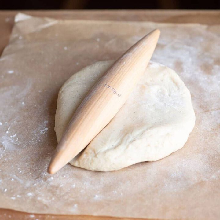 Holz-Nudelholz in einer konischen Form liegt auf einem St&uuml;ck Teig auf Backpapier. Der Teig ist rund und bemehlt.