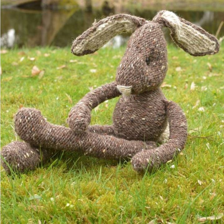 Gestrickter Hase aus brauner Wolle mit weißen Ohren sitzt auf grünem Gras vor unscharfem Hintergrund.