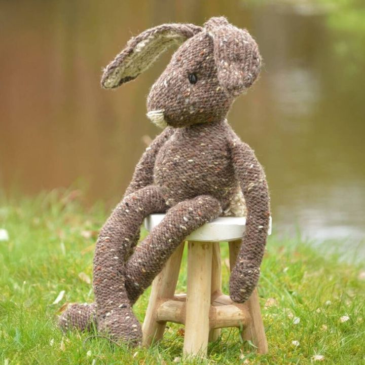 Gestrickter Hase aus brauner Wolle mit wei&szlig;en Ohren sitzt auf gr&uuml;nem Gras vor unscharfem Hintergrund.