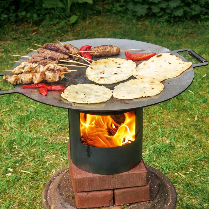 Ein Grill mit einer großen Metallplatte auf Holzblöcken. Oben Fleischspieße, Fladenbrote und Paprikastücke. Darunter brennt ein starkes Feuer. Der Grill steht auf einer Rasenfläche im Freien.