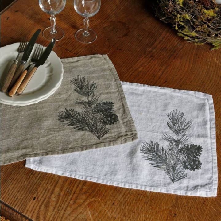 Zwei Leinentischsets, eines beige, eines weiß, mit Tannenzweig-Design auf Holztisch. Darauf ein Teller mit Besteck und zwei leere Weingläser.