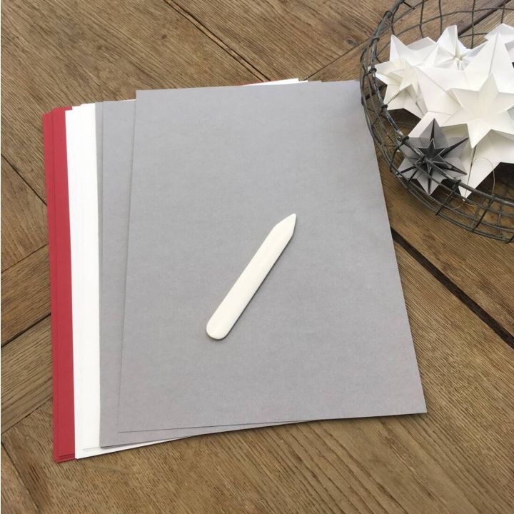 Mehrere B&ouml;gen farbiges Papier in Graut&ouml;nen liegen auf einem Holztisch mit einem wei&szlig;en Falzbein darauf; rechts ein Korb mit gefalteten Papiersternen.