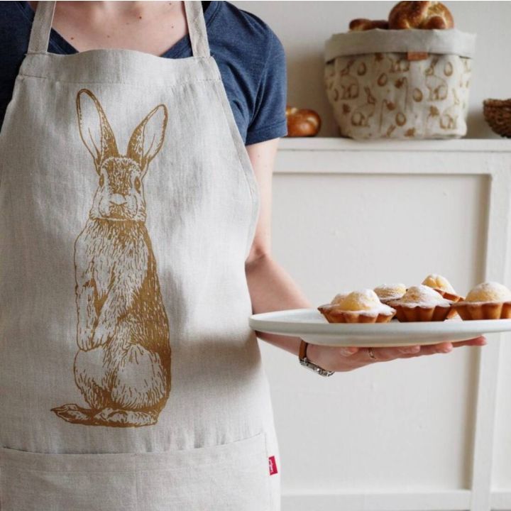 Person in grauem T-Shirt trägt eine helle Schürze mit aufgedrucktem Hasenmotiv. In der rechten Hand hält sie einen Teller mit sechs kleinen, mit Puderzucker bestreuten Muffins. Hintergrund: hell, mit dekorativem Brotkorb.