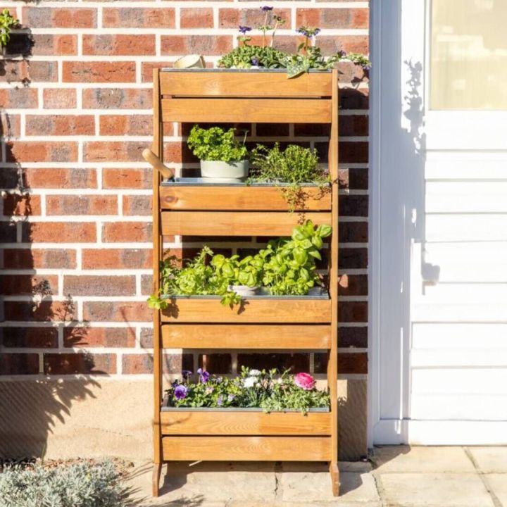 Vertikaler Holzgarten mit vier Ebenen, gefüllt mit verschiedenen Pflanzen und Kräutern, vor einer Ziegelwand. Der Pflanzenständer enthält Basilikum, Petersilie, Thymian und blühende Pflanzen. Teilweise beschattete Fläche neben einer weißen Tür.
