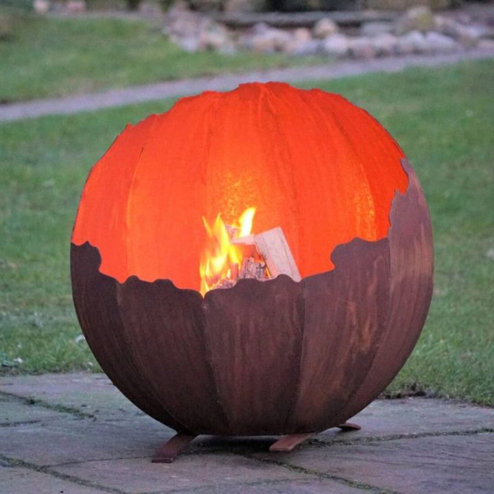 Kugelförmige Feuerschale aus Metall mit unregelmäßig geschnittenem Rand, in der ein Feuer brennt. Sie steht auf einer Terrasse mit Gras im Hintergrund.