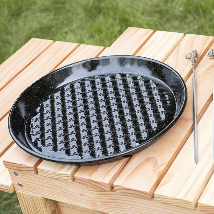 Schwarze, runde Grillpfanne mit Noppenstruktur, liegt auf einem hellen Holztisch im Freien; rechts daneben ein langer Metallspie&szlig; mit Haken am oberen Ende.