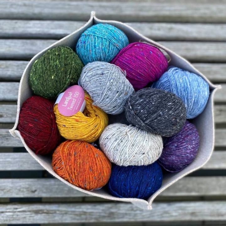 Zwölf bunte Wollknäuel in einem quadratischen Stoffkorb, auf einer Holzlatte sitzend. Labels teils sichtbar.