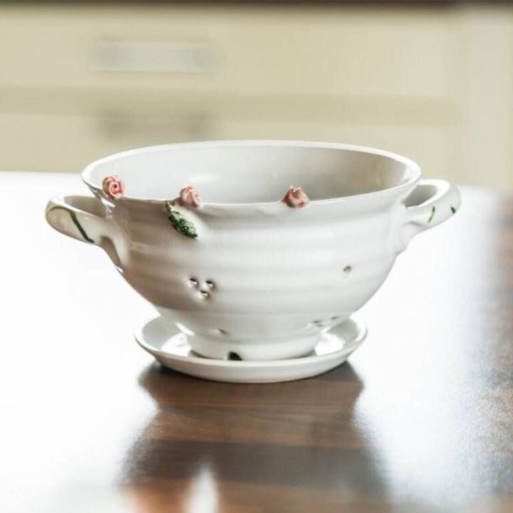 Weiße Keramik-Schale mit zwei Henkeln, floralen Verzierungen und kleinen Löchern, auf passendem Unterteller stehend; geeignet als dekoratives Sieb oder Obstschale.