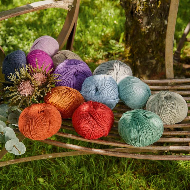 Mehrfarbige Wollknäuel auf einem Metallstuhl im Freien. Die Farben sind Orange, Rot, Blau, Grün, Lila und Grau. Links liegen dekorative Pflanzen, der Hintergrund zeigt Gras und einen Baumstamm.