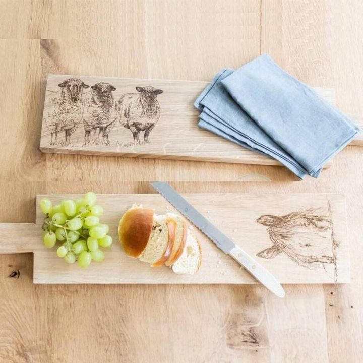Zwei Holzschneidebretter mit gravierten Schafmotiven auf einem Holztisch. Eines hat ein blaues Stoffservietten-Set darauf, das andere zeigt Trauben, geschnittenes Brot und ein Messer.