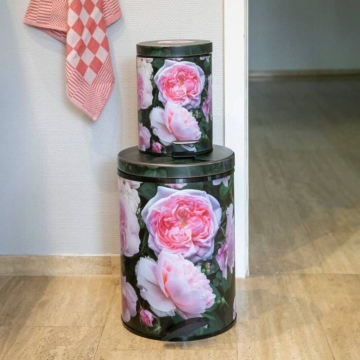 Zwei runde Mülleimer in unterschiedlicher Größe mit rosafarbenem Blumenmuster auf dunklem Hintergrund stehen übereinander auf einem hellen Boden vor einer weißen Wand; links hängt ein rot-weiß kariertes Geschirrtuch.